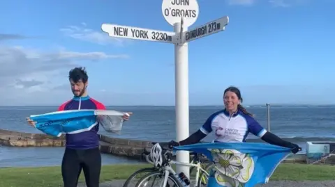 JES OAKLEY-STAFFORD Photo of Jes Oakley holding a Yorkshire flag, standing next to her husband at the John O'Groats sign, overlooking the sea.