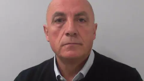 A bald man wearing a blue shirt with a black jumper on top. It is a police mugshot. 