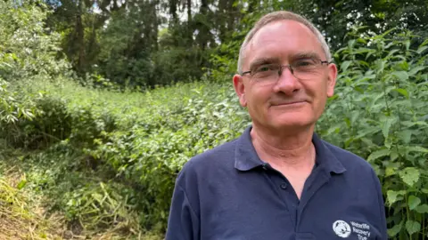 A man with short grey hair and glasses, wearing a dark blue polo shirt with a white logo on it. He is smiling at the camera while stood in front of a large stinging nettle bush