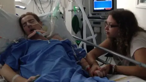 Matt Lodge in a hospital bed with his partner at his bedside