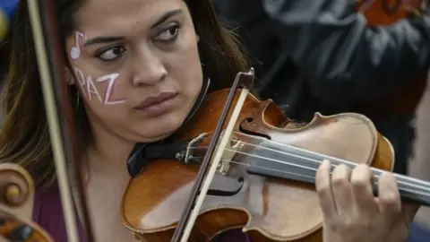 Getty Images A musician of the conservatory of the National University of Colombia plays the violin with her face painted that reads "peace" during protests and a national strike against the government of President Ivan Duque on November 27, 2019 in Bogota, Colombia