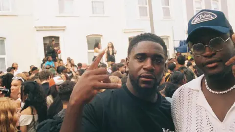 Kareem wears a white shirt, a blue cap and metal-rimmed glasses. His brother standing beside him wears a black T-shirt. He is holding his hand up with his index and little finger sticking out. Behind them a large crowd stand in the street. 