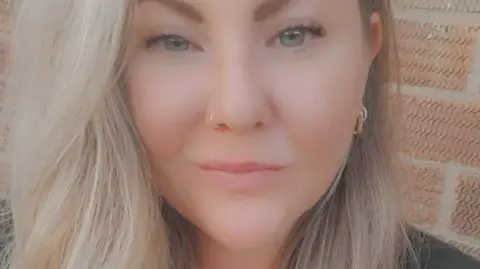 Kayleigh Glendon Kayleigh, a woman in her 30s with long blonde hair, standing in front of a red brick building 