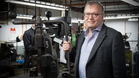A man wearing glasses, a grey blazer and a light blue shirt stands beside a heavy iron fly press in an industrial workshop. Behind him, strip lighting illuminates a cluttered workspace filled with machinery, tools and equipment. A second worker can be seen in the background on the left of the frame.