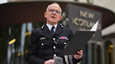 PA Media A balding middle aged man wearing police uniform stands outside New Scotland Yard police headquarters. He is reading a statement from a clipboard which he is holding in his left hand. 
