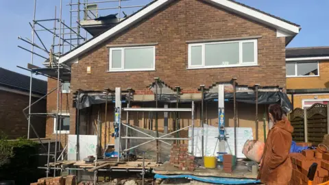 Simon Thake A woman looks side on at the back of a house that appears to be propped up with steel beams