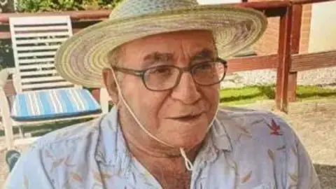 An elderly man wearing glasses and a sunhat, while wearing a white shirt. He is sat in a chair outside looking off to the right of the camera