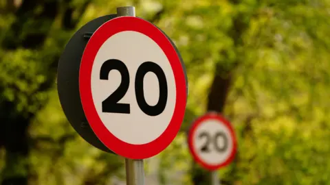 A generic image of a red and white 20mph speed limit sign on a blurry tree background. A second sign, slightly blurred, can be seen behind the first, more prominent sign.