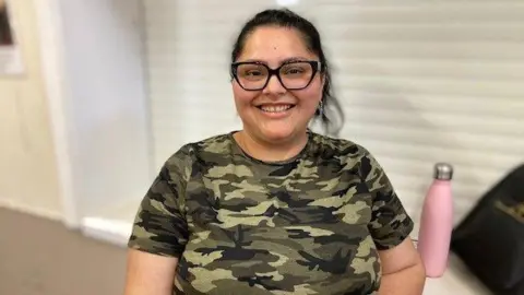 A woman stands in a room. She has straight black hair glasses, and she is wearing a t-shirt with a camouflage pattern. A table with a pink water bottle can be seen behind her.