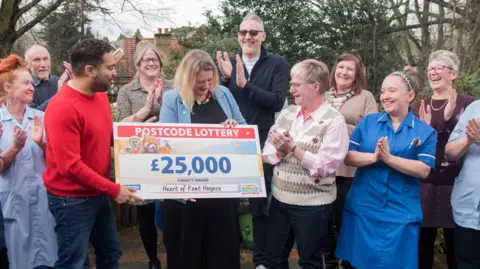 PEOPLE'S POSTCODE LOTTERY A group of people stand clapping as a large cheque for £25,000 from the People's Postcode Lottery is handed over to the Heart of Kent Hospice team. 