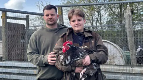 Tom, a man with short brown hair and brown top, with his son, also called Tom, who has brown hair, a brown jacket and blue top, holding a cockerel. There is a wooden and mesh cage behind them in an allotment and some trees.