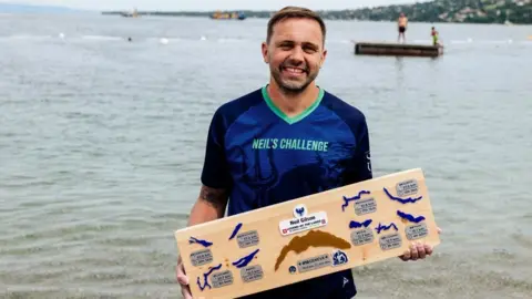 Neil Gilson is wearing a blue T-shirt with the words Neil's Challenge on it. He is standing in front of a lake holding a plaque showing all the lakes he has swum through.
