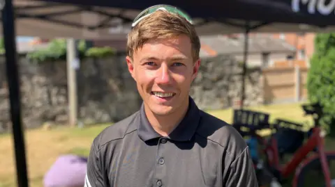 A brown-haired man with sunglasses on the top of his head, wearing a black polo shirt, standing in front of a black gazebo and red bike, with a grassy garden and stone wall in the background.