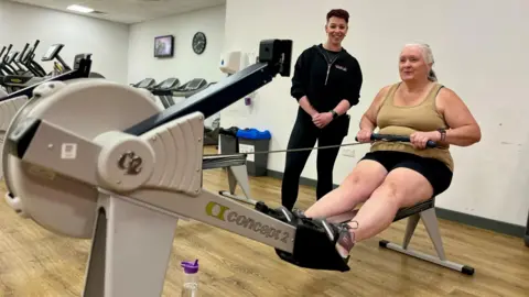 Nikki Fox/BBC Deborah Caitens-Smith on the rowing machine at Brandon Leisure Centre in Suffolk. She is with her personal trainer, who is standing to her right. Ms Caitens-Smith is wearing a khaki vest with black cycling shorts and trainers, there is a water bottle on the floor.