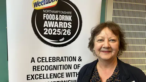 A woman in a black top stands next to a white board promoting the upcoming Northamptonshire Food and Drink Awards.