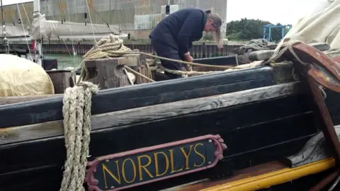 Jamie Niblock/BBC The side of the Nordlys ship is painted black with a sign that says the boat's name in gold. William Lund is standing on the boat's deck, bending down looking at rope.