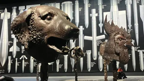 Photograph of the Ai Weiwei exhibition at Manchester's Aviva Studios. The image shows animal head sculptures and a large wall mural.