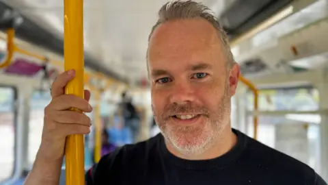 BBC Keith Watson, who has grey hair and a beard and wears a black T-shirt, standing in a train carriage holding onto a yellow pole.
