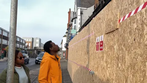 George Carden/BBC Simba, wearing a yellow rain coat, looks carefully at the remains of his burnt flat. There is a large plywood wall around the shop which is on the ground floor and there are burnt pieces of wood on top of the wall. 