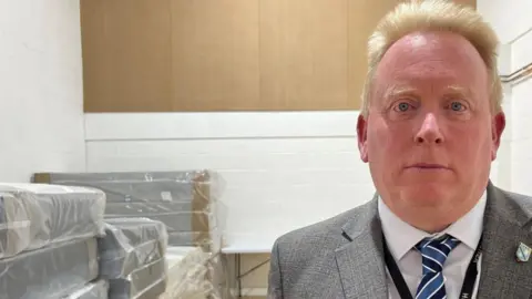 Mr Tilling standing in front of a pile of new mattresses each still covered in plastic wrapping. He has blond hair and is dressed in a shirt and tie with grey jacket.