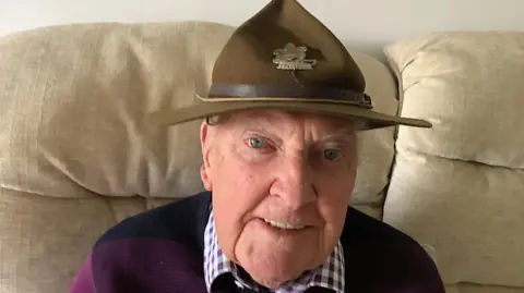 Geoff Pinney Head and shoulder shot of an older man sitting on a beige sofa wearing a checked shirt and burgundy sweater. He is wearing an old wide-brimmed scout hat with a gold emblem on the front.