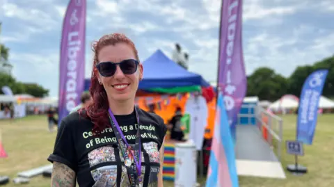 Kaye Nicholson smiling at the camera. She is wearing a black top with a purple lanyard in front of a blue tent which has two purple flags with the word 'liberate' on them.