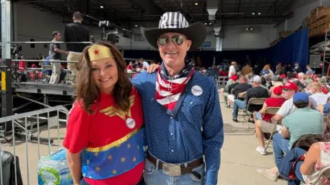 Maleesa Meyers, left, is wearing a red, white and blue Wonder Woman costume and Michael Milton, right, is wearing a black cowboy hat with Trump's name on the front and an American flag scarf along with a button-down long sleeve shift and thick leather belt. The two are photographed at the back of a JD Vance rally in an airplane hangar. 