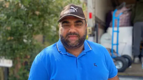 Sarmad Dar, wearing a blue t-shirt and brown Puma baseball cap, stands in front of a tree 