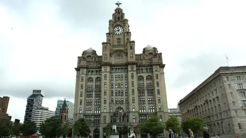 People walking past the Royal Liver Building by the docks in Liverpool on a dry day.