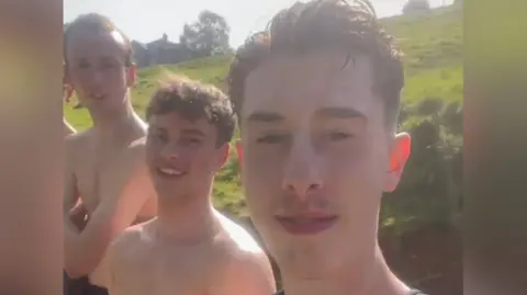 Three teenage boys stood outdoors at a swimming spot.