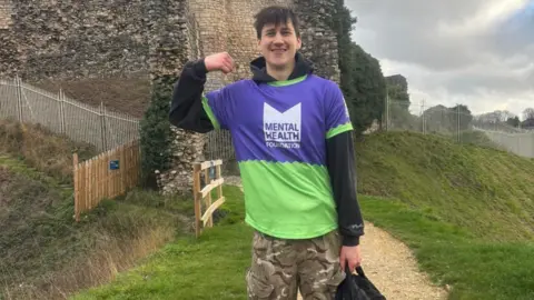 A young man wearing a black hoodie and camouflaged trousers holding a black backpack and wearing a purple and green top which says 'Mental Health Foundation' on it. He has short brown hair and is smiling with his fist clenched towards his face as he flexes his arm. He is stood on the top of a hill next to some brickwork.