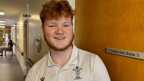 A teenage boy with curly red hair and a beard, wearing a white WRU branded polo shirt and smiling as he looks at the camera.