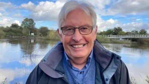 A man in a blue waterproof coat, blue sweater and blue shirt smiles at the camera. He is standing in front of a river, on the other side of the river is a line of trees