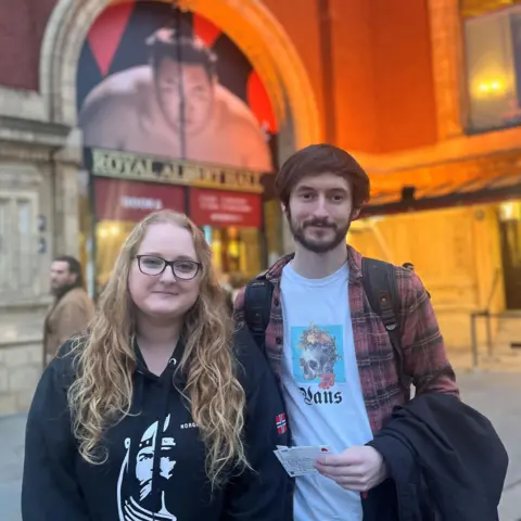 Flora Drury/BBC Sian, wearing a black top with long blonde hair and glasses, stands with Luke, wearing a plaid shirt and a skull t-shirt, in front of a picture above an entrance door showing a sumo wrestler staring into the camera