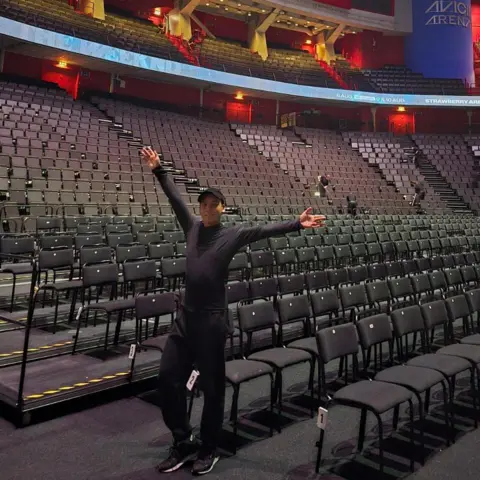 Supplied Hanyu Wong stood in an empty arena. He is wearing all black and his arms are aloft as he looks towards the camera.