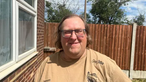 Alexander Stevanovic/BBC Jensen Parker wears a brown T-shirt, standing outside, with a white-framed window to his right, and a brown-painted garden fence behind him