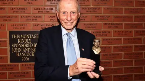 Port Vale FC John Rudge is in a suit, standing in front of a brick wall. Each brick has someone's name on it. He is holding a small bronze statue which is a bust of himself