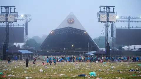 Ben Birchall/PA Media The Pyramid Stage in the distance and a large crowd of volunteers litter picking at Glastonbury. There is litter all over the field.