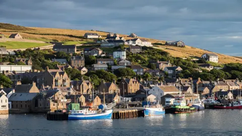Getty Images Stromness from the harbour. It is sunny though the sky behind the hill is dark. There are boats in the harbour.