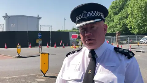Ch Insp Iain Wyke is wearing a white police uniform shirt with a black tie and a police hat. He is stood at the side of a road at Sefton Park opposite fencing and the back of a large stage. A number of red and white cones and fencing green and metal fencing is in the background.