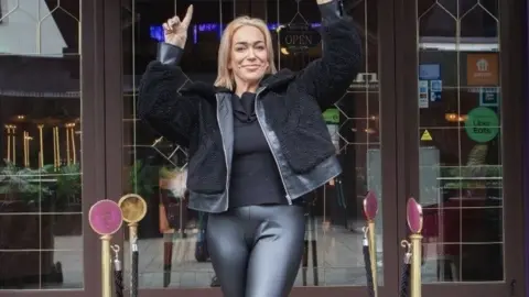 Melanie Prince stands outside a bar on a red carpet. She is holding up her hands. 