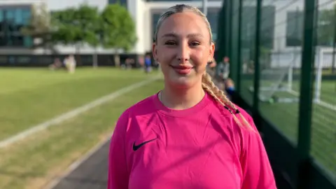 A woman with long, tied-back blonde hair smiles and a pink shirt, smiles as she stands at the side of a football pitch.