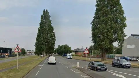 A main road with vehicles travelling in both direction. A couple of road signs and large trees line each side of the road.