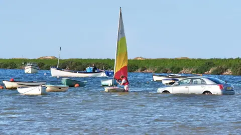 GaryPearson Photography A man is standing in the water with a llife jacket on adjusting a brightly-coloured striped sail to a small boat. Boats are bobbing in the water around him and a silver saloon-type car is submerged in the water.