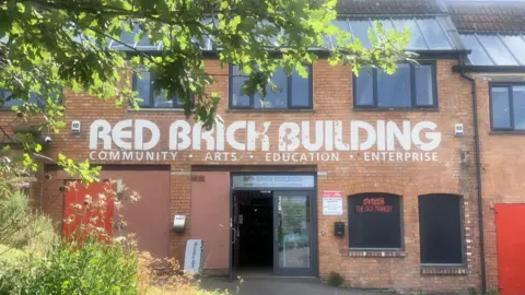 Red Brick Building The outside of the Red Brick Building entrance with its name above the door and some foliage on the left foreground