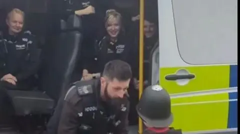 Durham Police Tyler wearing a police helmet and pictured from the back meeting a police officer. They are standing in front of a police van with other officers watching them.