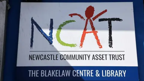 A sign with NCAT in different school artwork-style colours. The A has been made into a person. Underneath it reads Newcastle Community Asset Trust. The last lines says The Blakelaw Centre and Library.