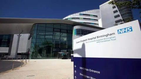 An exterior view of the Queen Elizabeth hospital showing a large building with glass frontage leading to its reception area
