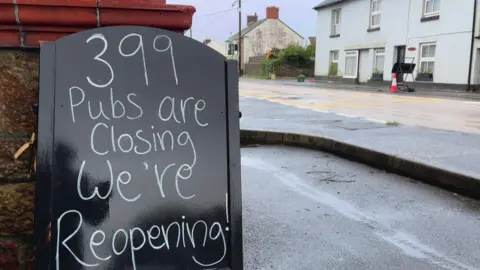 A pub sign next to a busy main road - It reads "399 pubs are closing , we're reopening !"