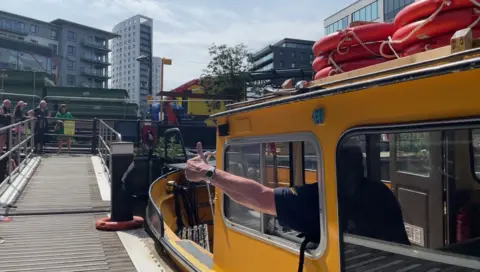 A bright yellow water taxi in Leeds. A man has his arm out of the window of the boat giving a thumbs up sign towards a group of onlookers standing at the entrance to a jetty. Office and apartment tower blocks can be scene in the background against a cloudy sky.
